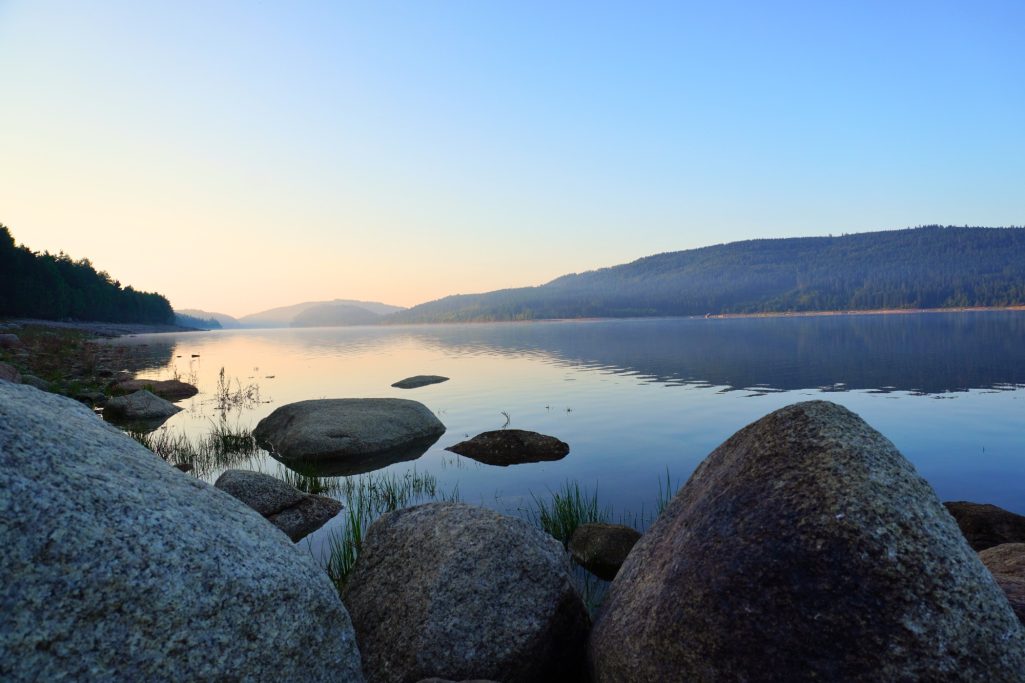 Ruhiger See mit Steinen im Vordergrund und sanften Hügeln im Hintergrund bei Sonnenaufgang.