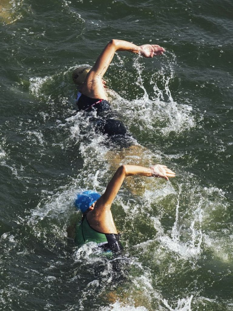 Zwei Schwimmerinnen, eine mit blauer Badekappe, schwimmen im offenen Wasser.