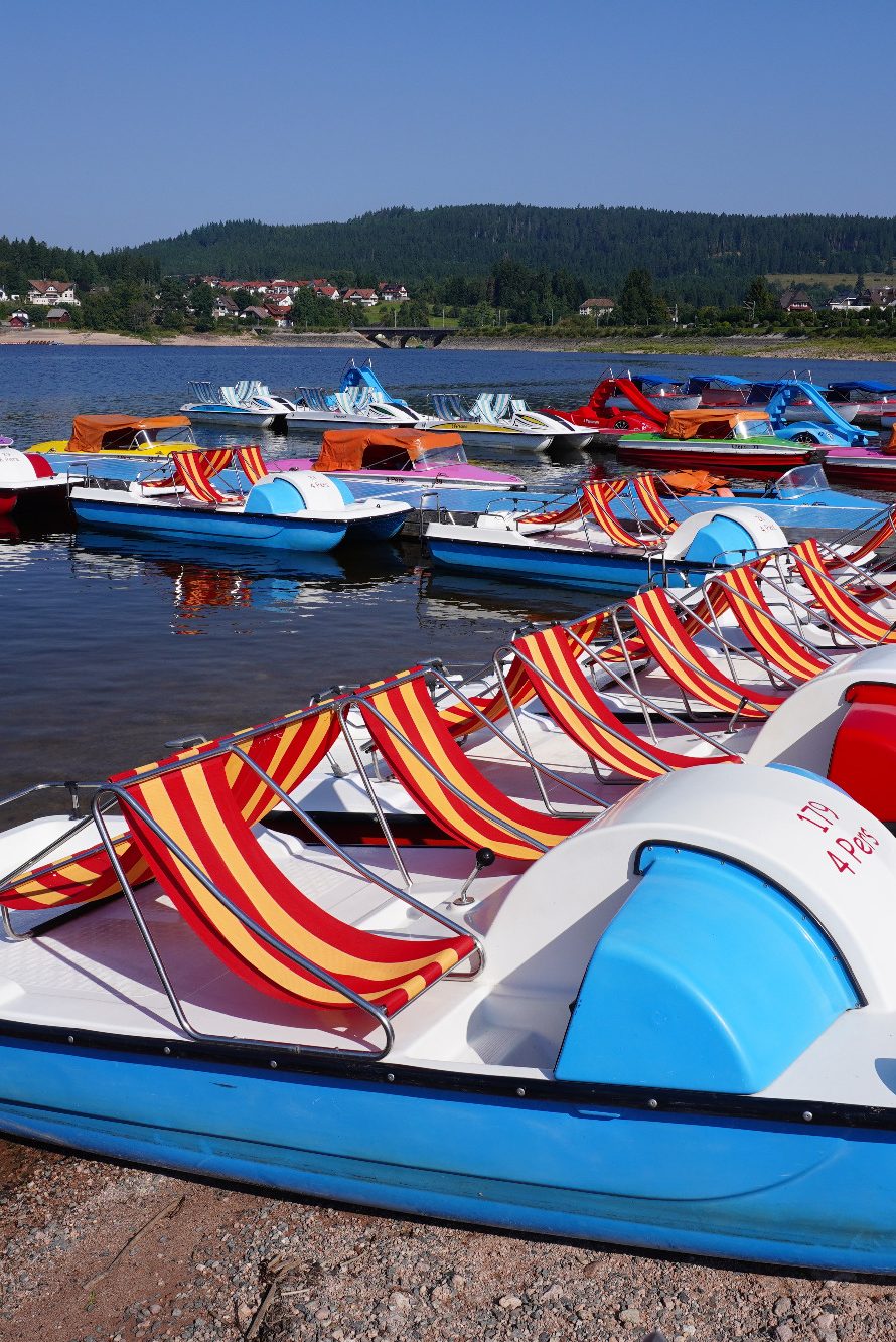 Bunte Tretboote am Ufer eines ruhigen Gewässers mit Hügeln im Hintergrund.