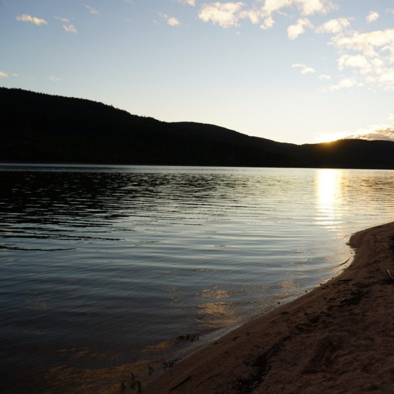 Ruhiger See bei Sonnenuntergang, umgeben von sanften Hügeln und leicht bewölktem Himmel.