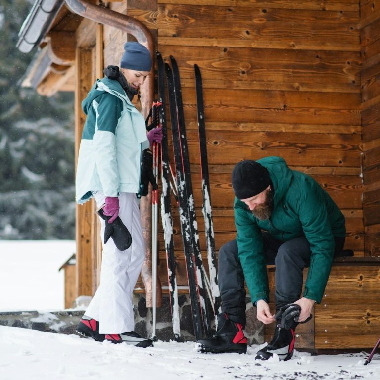 Langlauf Zwei Personen schnüren die Schuhe vor einer Holzhütte im Schnee, um Ski zu fahren.