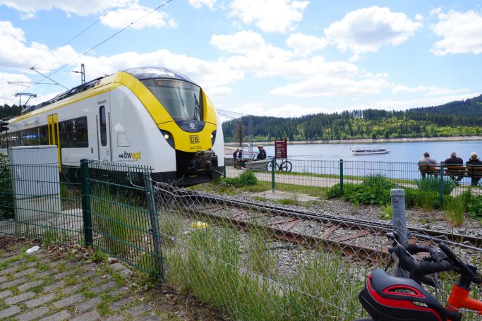 Modern Zug an einem See mit Passagieren auf dem Bahnsteig und grüner Umgebung.
