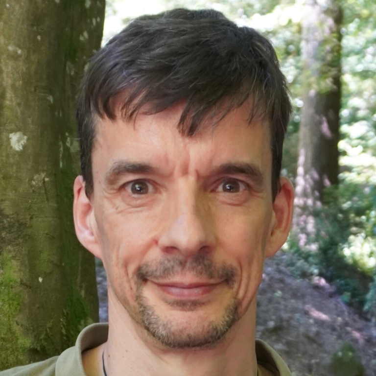 Porträt eines Mannes im Freien, umgeben von Bäumen. Freundlicher Gesichtsausdruck.