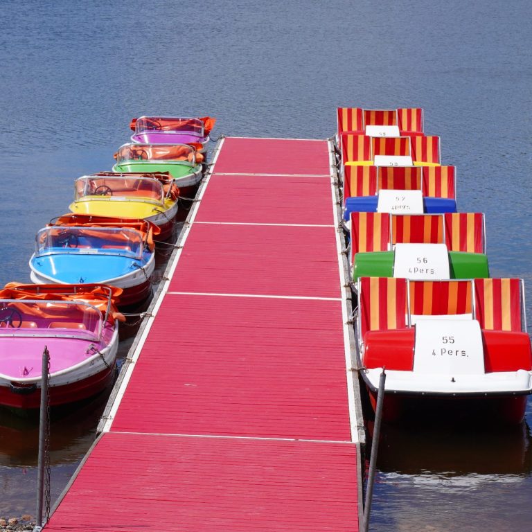 Bunte Tretboote auf einem Steg am Wasser, mit roten und gestreiften Sitzen.