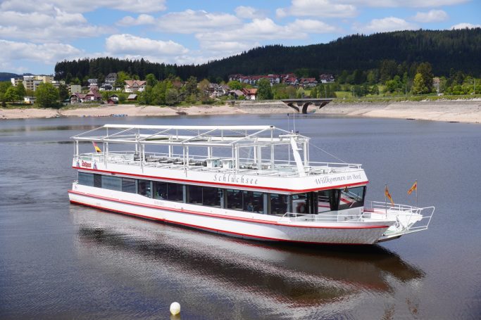 Weißes Ausflugsboot mit rotem Streifen auf ruhigem Fluss, umgeben von grüner Landschaft.