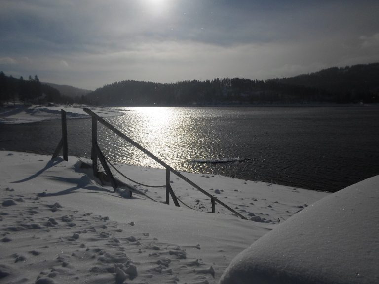 Schluchsee im Winter Schneebedeckte Landschaft mit einem See und einer reflektierenden Sonne im Hintergrund.
