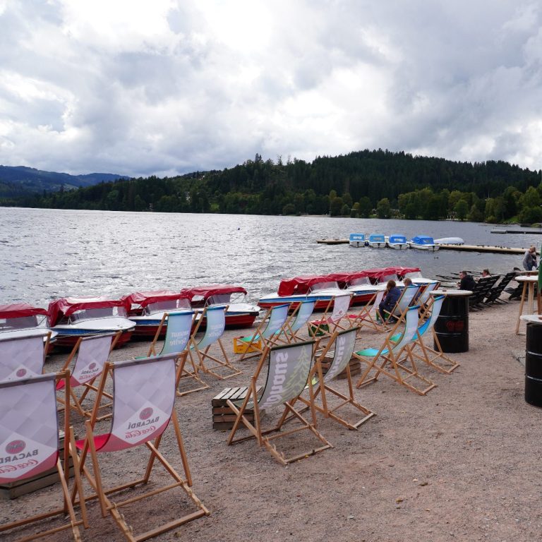 Blick auf einen See mit Liegestühlen am Ufer und bewaldeten Hügeln im Hintergrund.