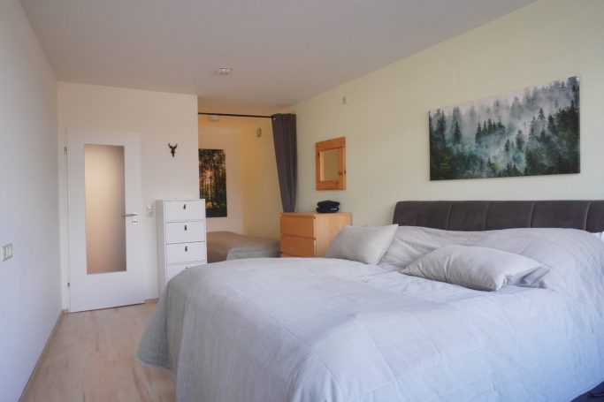 Helles Schlafzimmer mit großem Bett, Kommode und einem gemalten Landschaftsbild an der Wand.
